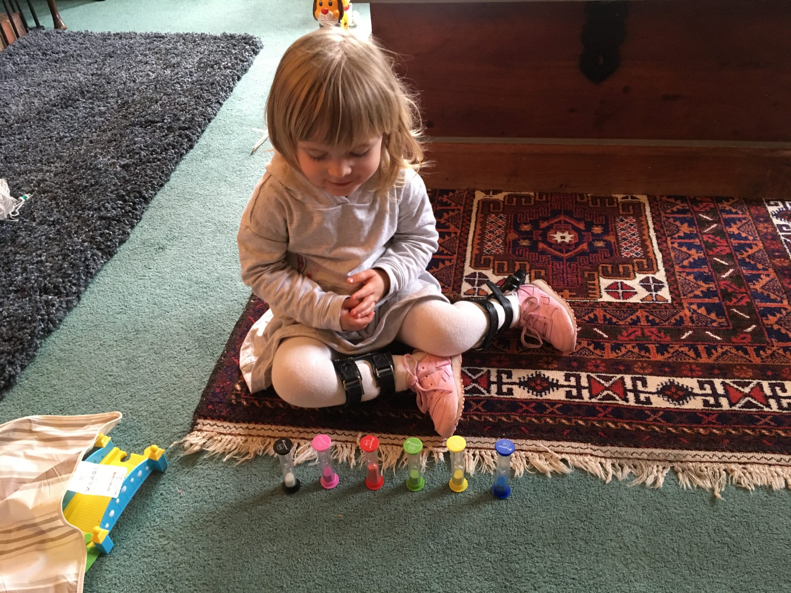 A young girl with club foot sits on a patterned carpet, playing with some colourful egg timers.