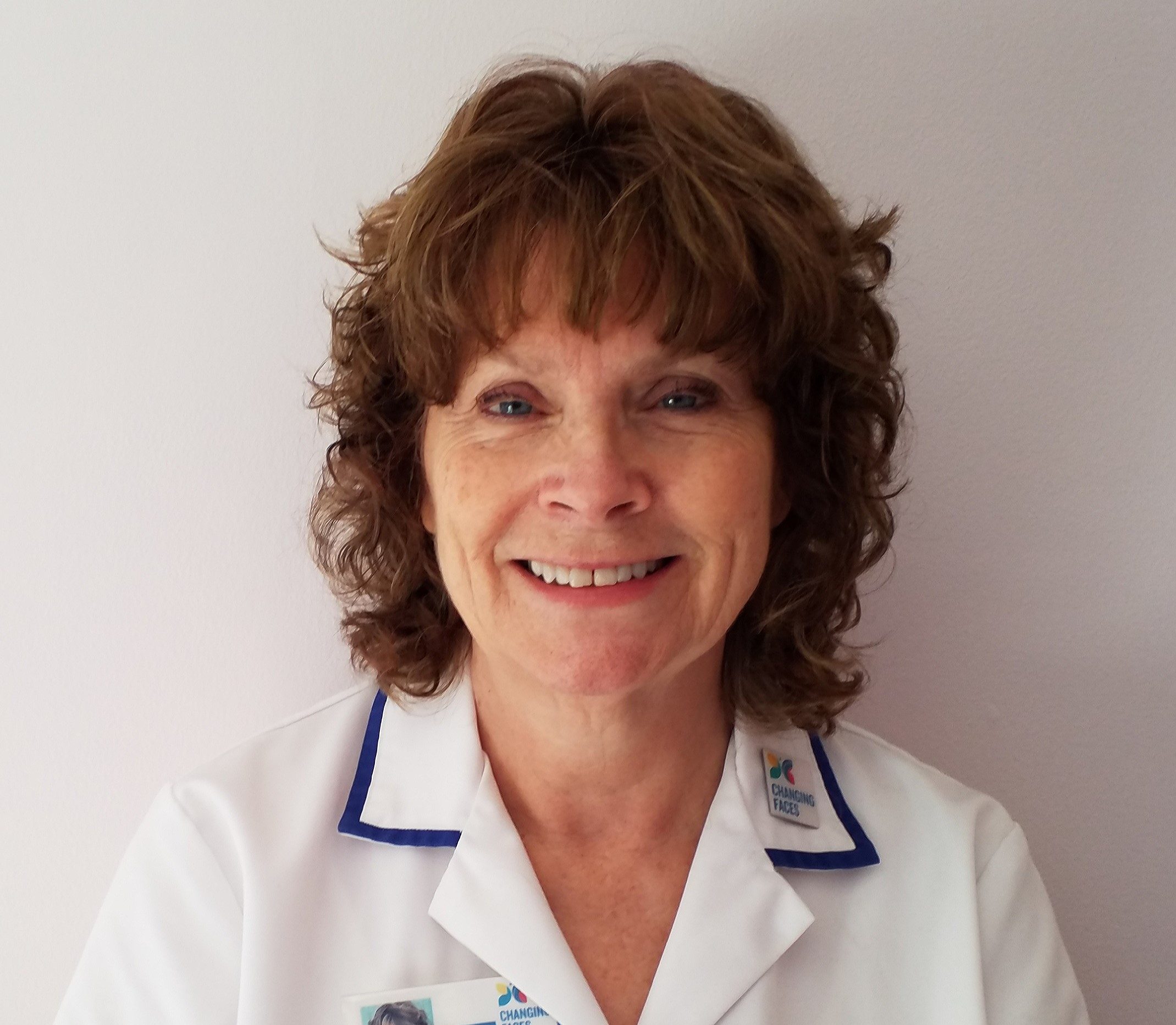 A skin camouflage practitioner wearing a white medical coat with a Changing Faces badge, smiles at the camera. She has short curly hair.