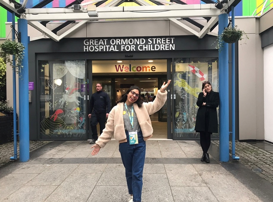 A young woman in a white jacket raises her arms and smiles in front of Great Ormond Street Hospital.