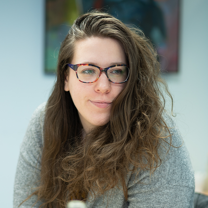 A young woman with curly brown hair, wearing glasses and a grey cardigan, looks to the right of the camera, slightly smiling.