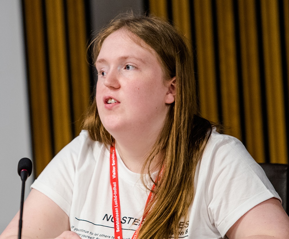 A teenage girl who has a facial cleft sits at a desk in front of a microphone. She has long hair and is speaking towards the left of the camera.