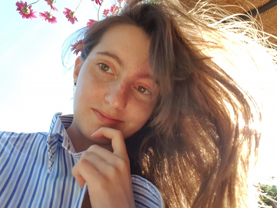 A girl with long brown hair looks down at the camera; behind her is a blue sky and pink flowers.