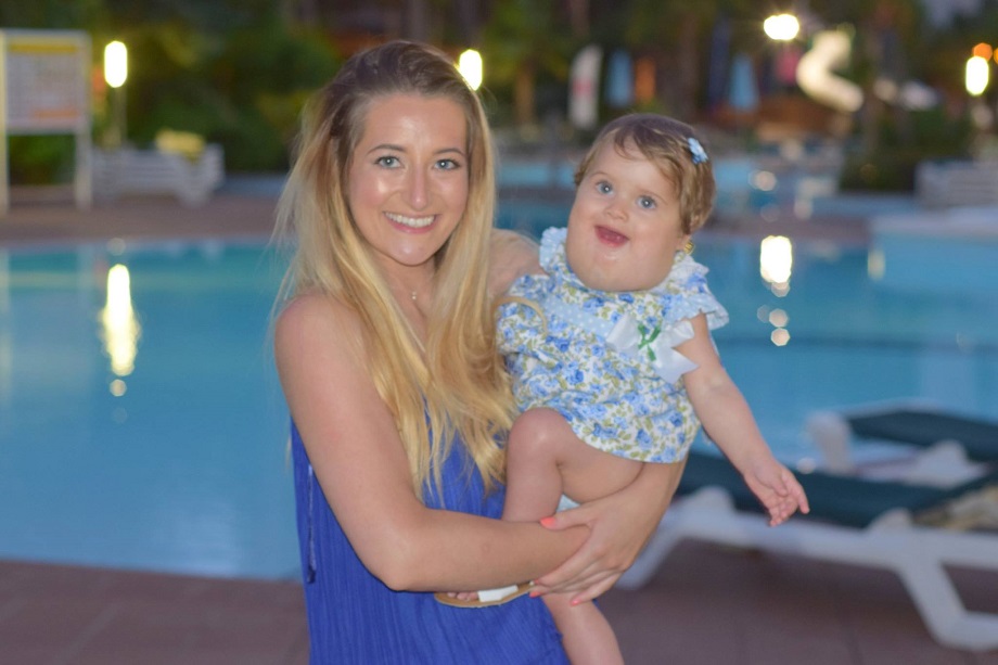 A young woman with long blonde hair holds her baby girl, who has a lymphatic malformation; behind them is a swimming pool.