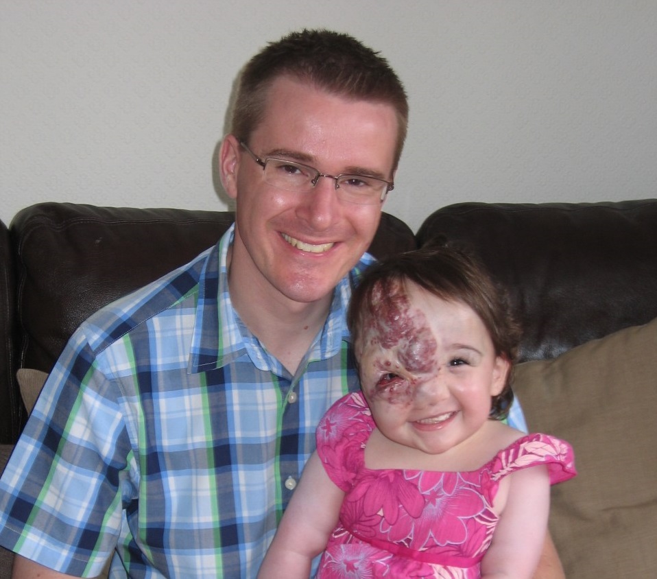 A man in a checked shirt holds his baby daughter, dressed in pink, on his lap. Both are grinning.