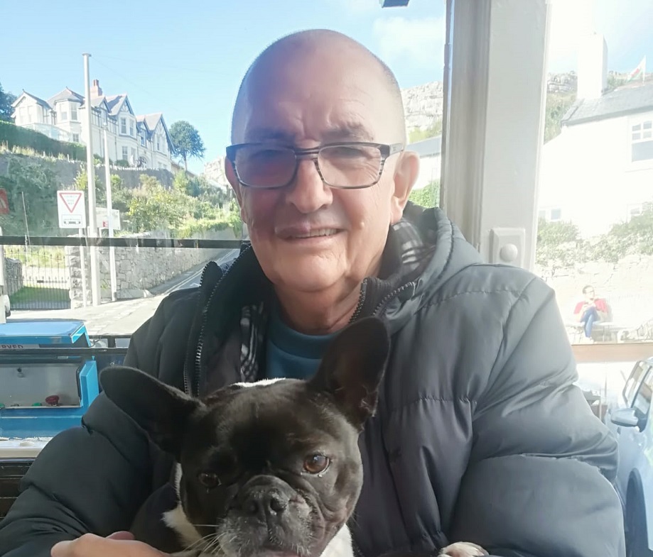 A man wearing glasses holds a small dog in his lap. Behind him is a window looking onto a sunny street.
