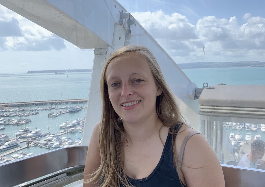 A young woman with long blonde hair sits in a Ferris wheel; below are boats in a sunny harbour.