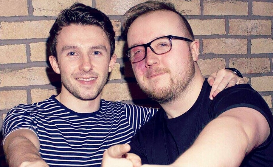 A man with a port wine stain birthmark on his face smiles next to another man, who's wearing a striped top. Both are smiling.