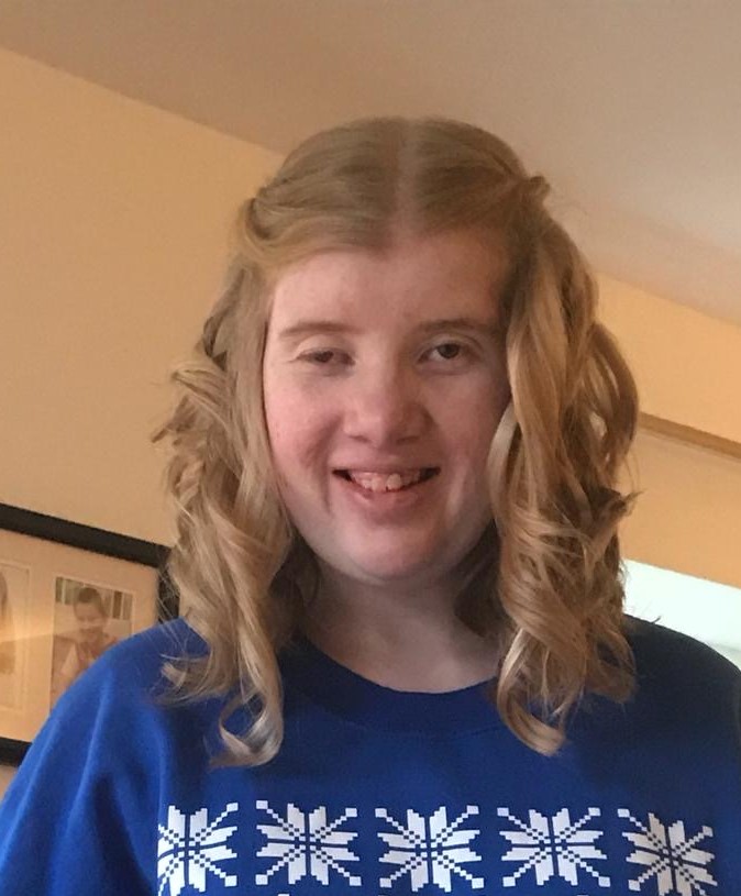 A young girl with ptosis wears a blue and white top and smiles at the camera. She has blonde curly hair.