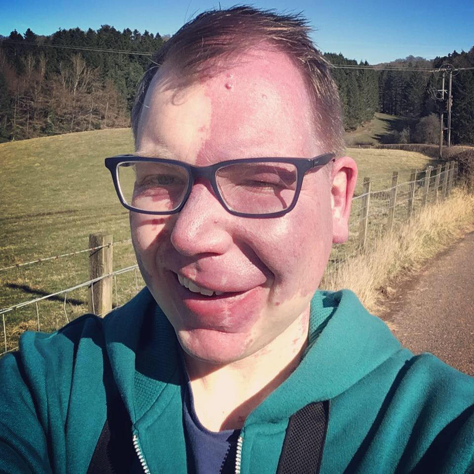 A man wearing glasses and a green hoodie takes a selfie on a country road. He is smiling.