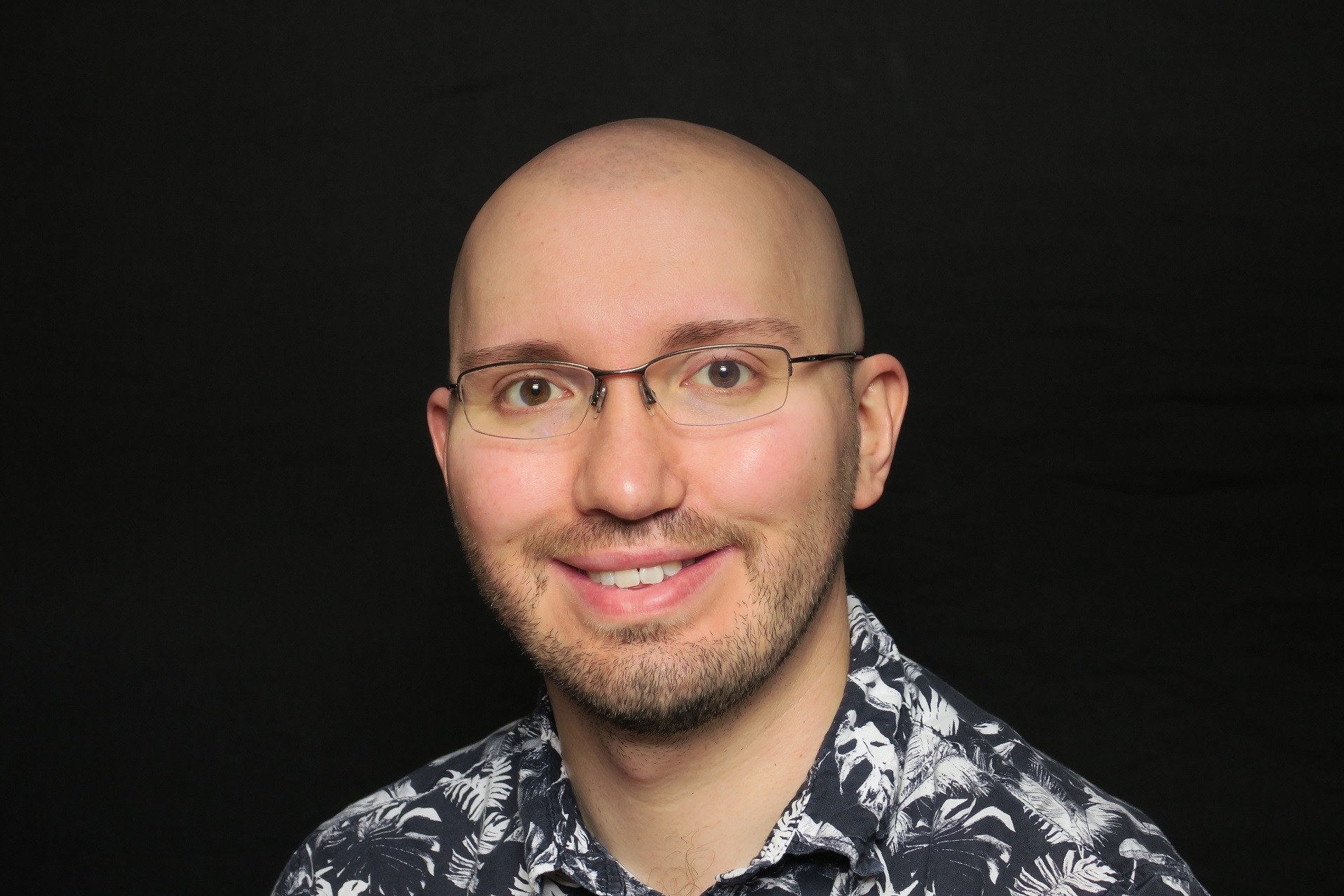 A man wearing glasses and a patterned black and white top smiles at the camera against a black background.