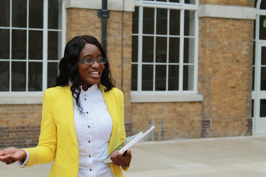 A woman wearing glasses and a bright yellow blazer holds a book and smiles to the right of the camera.