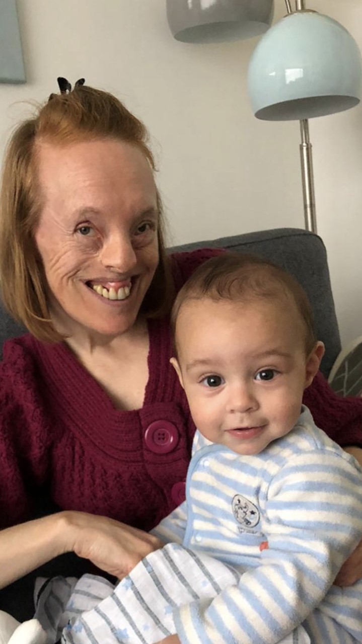 A woman wearing a red jumper holds a baby in striped pajamas. They're both smiling.