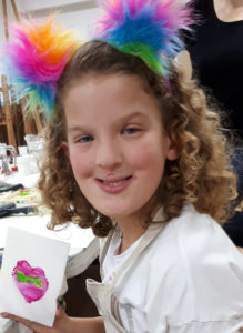 A photo of a young girl with curly brown hair, sitting at a table, holding a card she's made