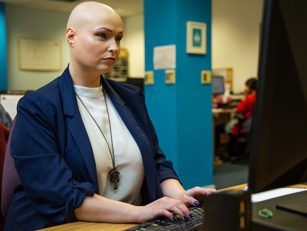 Brenda who has alopecia and wears a blue blazer and white top sits typing at a computer