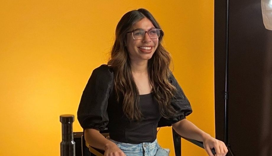 A woman in her early 20s, wearing a black blouse and blue jeans. She's sitting in a chair and there's a yellow back drop behind her. She has long brown hair and glasses.