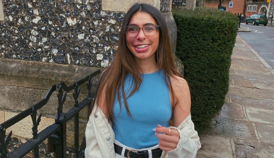 A woman in her early 20s, long dark hair, wearing glasses, a blue sleeveless t-shirt, washed out blue jeans with a black belt, and an off white cardigan. She's standing in front of a black fence, behind which is a stone building and a green bush.