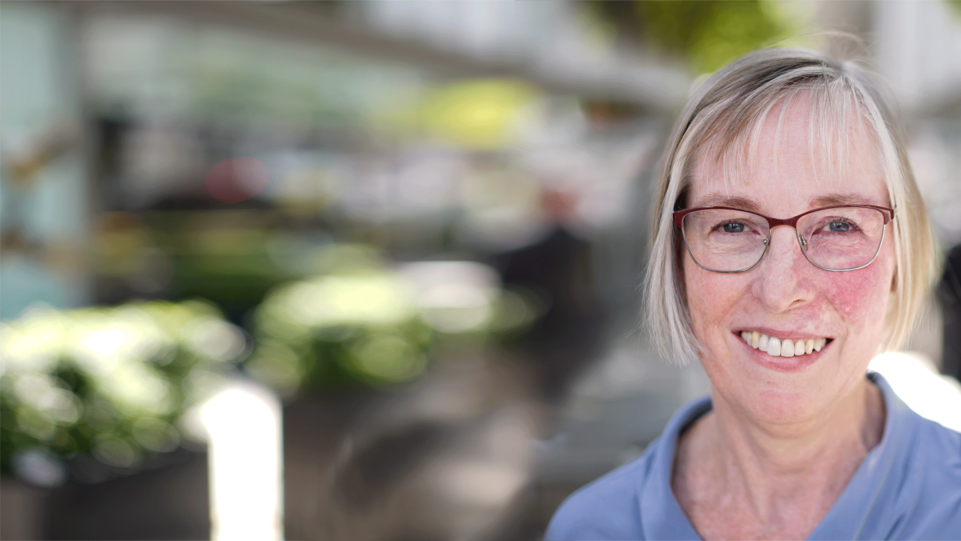 Susan wears glasses and a lilac top and stands outside on a sunny day. She has a mark on her cheek.