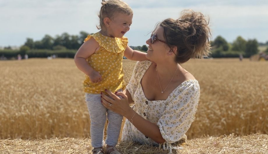 A woman wearing a white floral blouse and sunglasses. She is holding her daughter who's wearing a yellow top and white leggings. She is smiling at her mother. A field of straw is in the background.