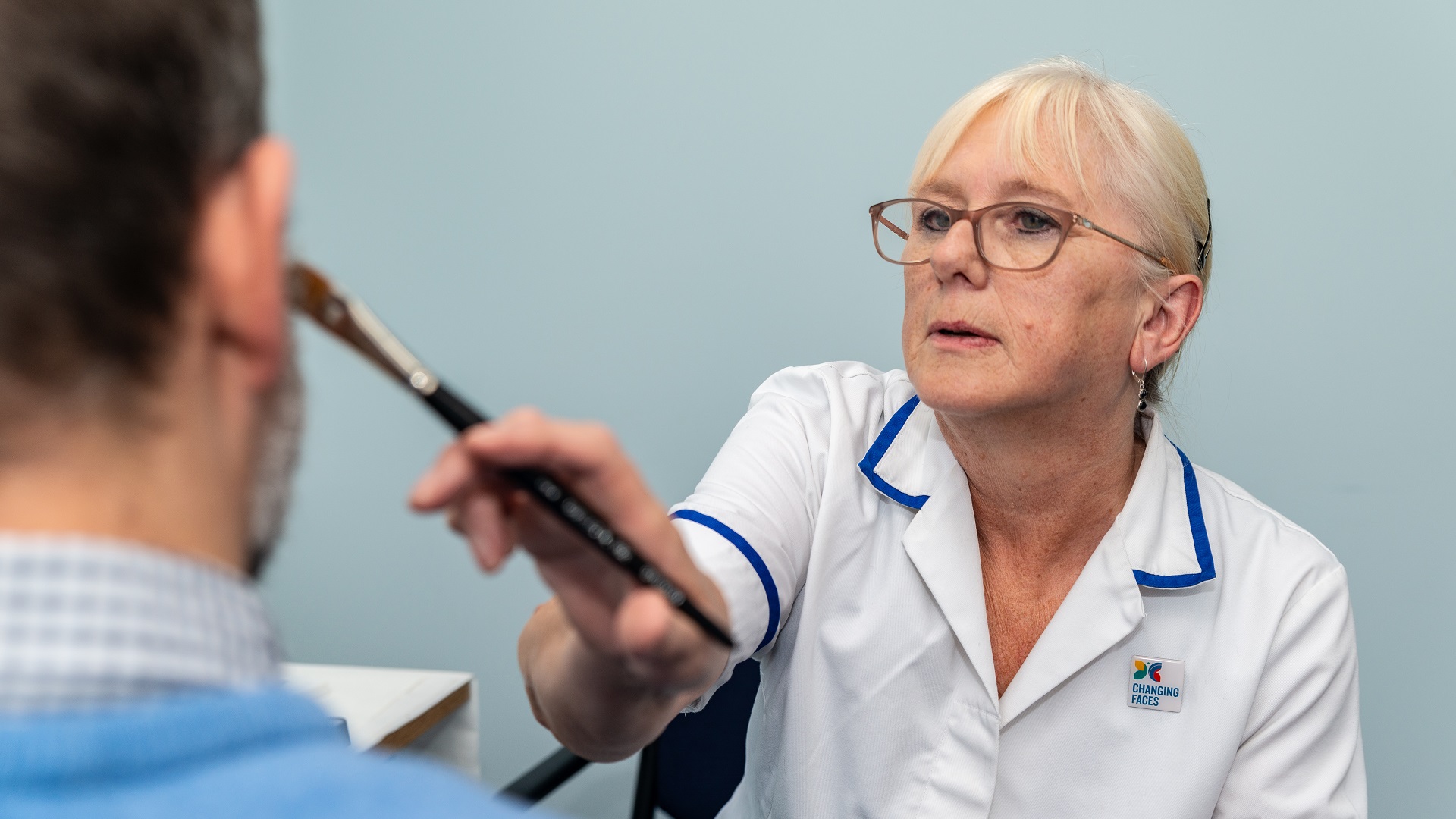 A woman in a nurse's top, wearing glasses with white hair tied up, applies skin camouflage products to a man's face. We can see the back of the man's head only - he has some stubble on his face and wears a shirt and a blue jumper.