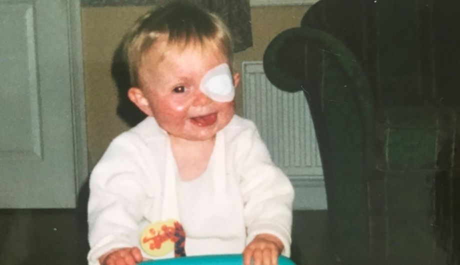 A toddler in a white cardigan with short blond hair and a eye patch pushes a trolley of toys