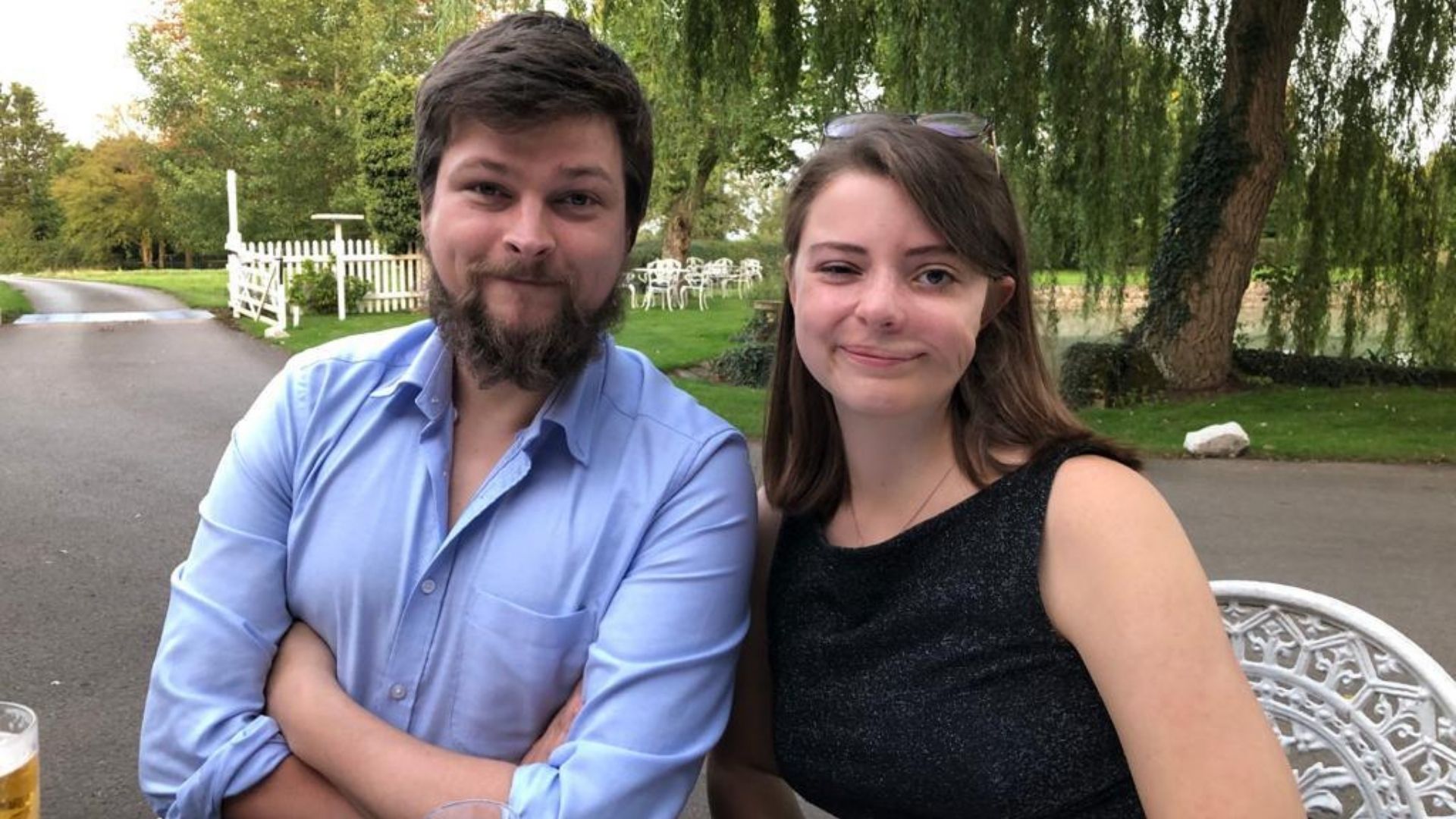 Bethan, a woman in her 20s, wearing a black blouse, with sunglasses in her hair, smiling at camera. Next to her is a man, 20s, with a bead, wearing a blue shirt. Behind them is a willow tree, a driveway, white gate and grass.