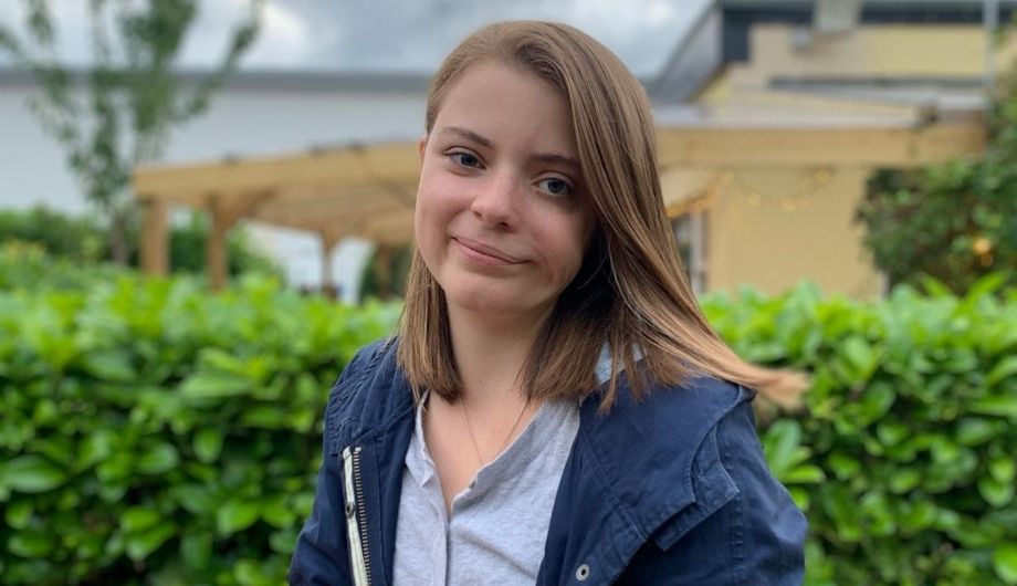 A woman, early 20s, shoulder length brown hair, blue jacket and blue shirt, sitting in front of a hedge.