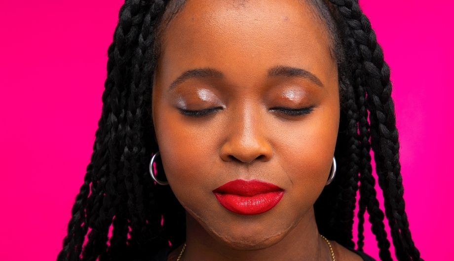 A young Black woman, 20s, she has her eyes closed and is wearing red lipstick and earrings. Behind her is a pink backdrop.