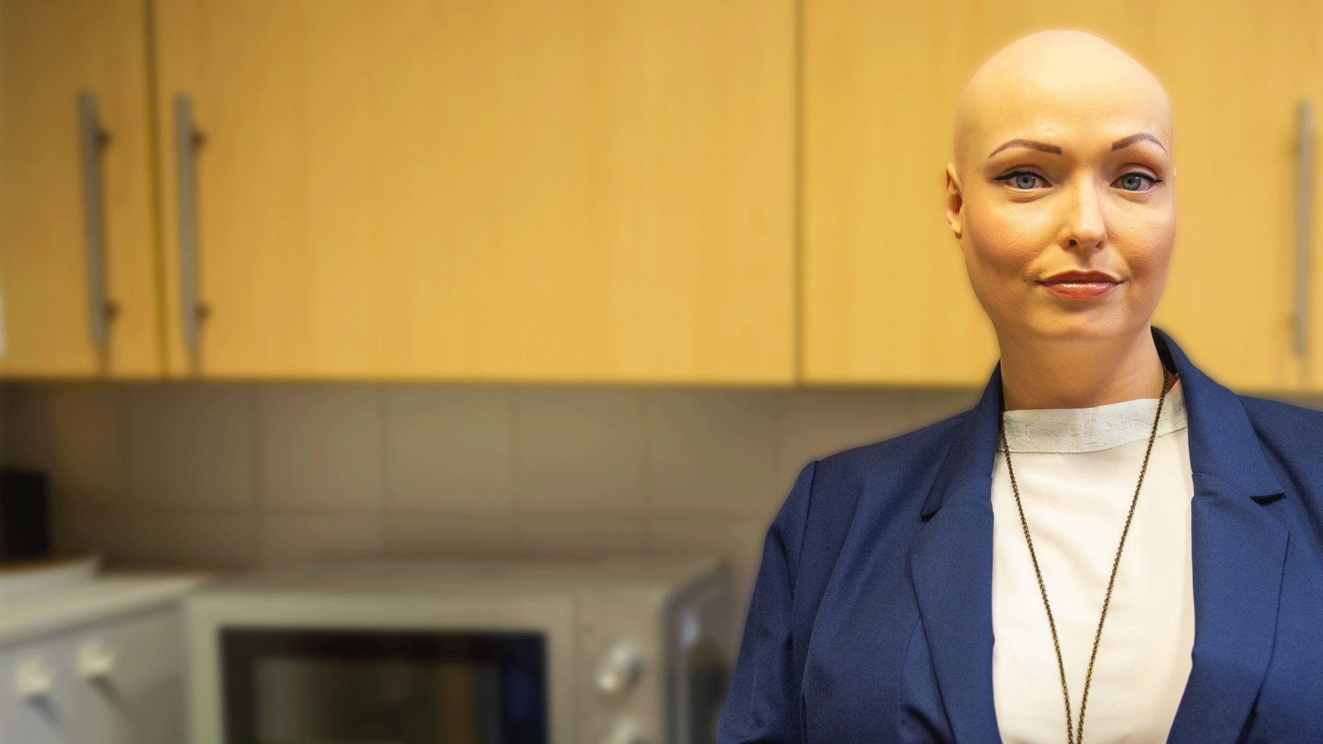 A woman with alopecia stands in the kitchen, gazing at the camera