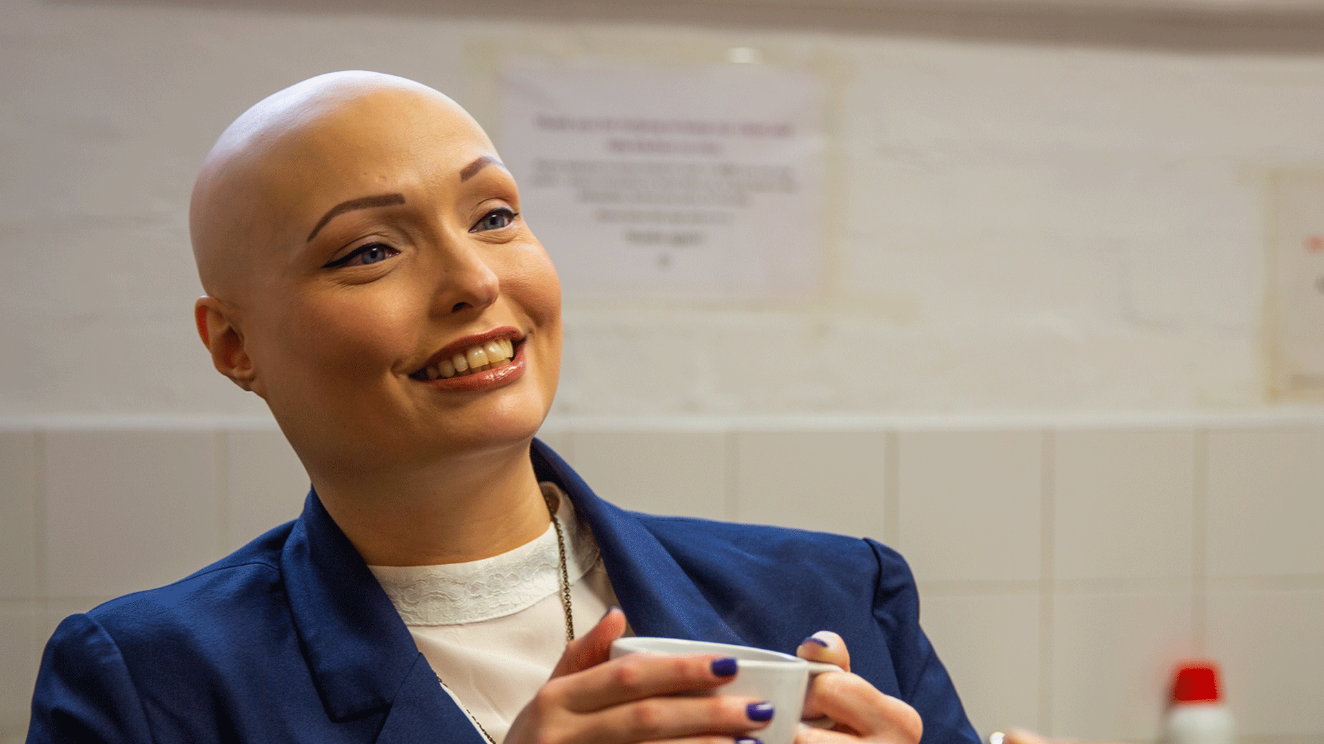 A woman with alopecia and holding a cup, smiles to someone off camera
