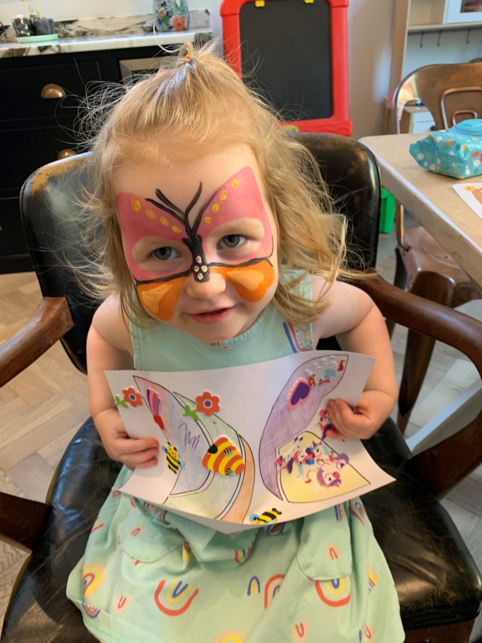 A small girl wearing butterfly face paint and holding a drawing of a butterfly