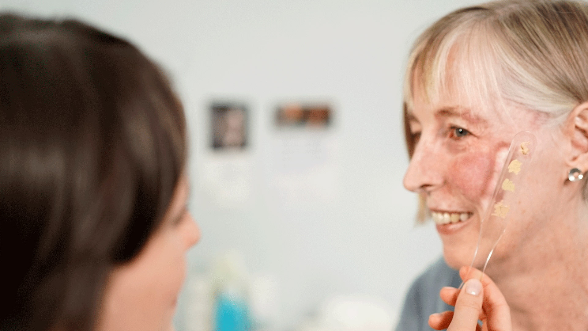 A woman holding a range of skin camouflage products up to the face of a white woman who has a facial birthmark.