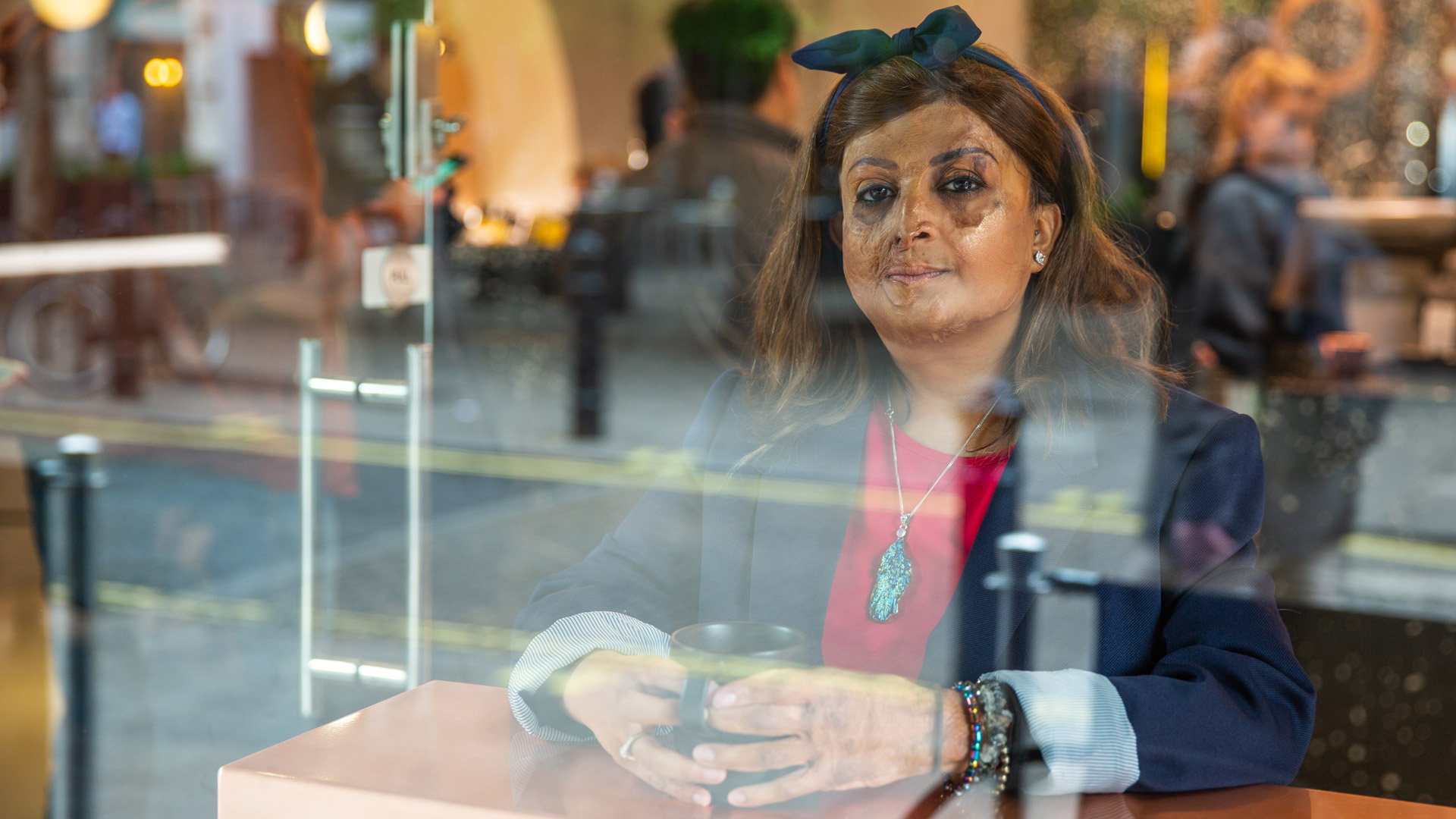 Tulsi sitting in the window seat of a coffee shop with a mug, and gazing to camera