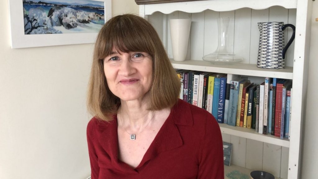 Heather Blake, a woman with shoulder length brown hair, wearing a red blouse, standing in front of a bookcase, filled with books and ceramics.