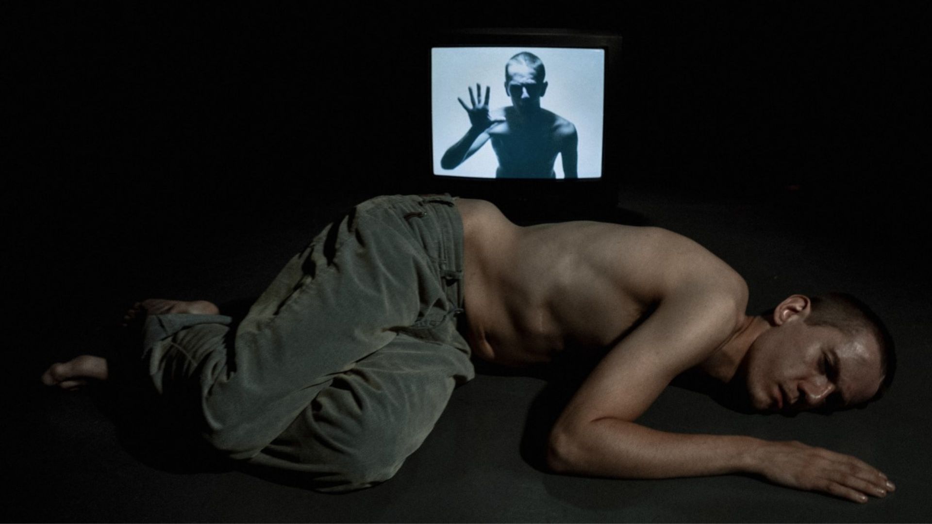 Robert, a young man in his 20s, topless wearing green trousers, lying on the ground. Behind him is a tv with an image of himself on the screen.