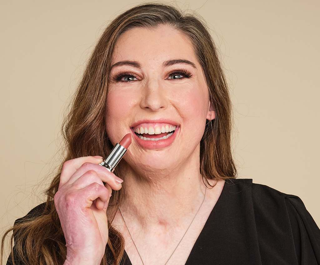 A woman in a black top smiles at the camera and holds up a lipstick.