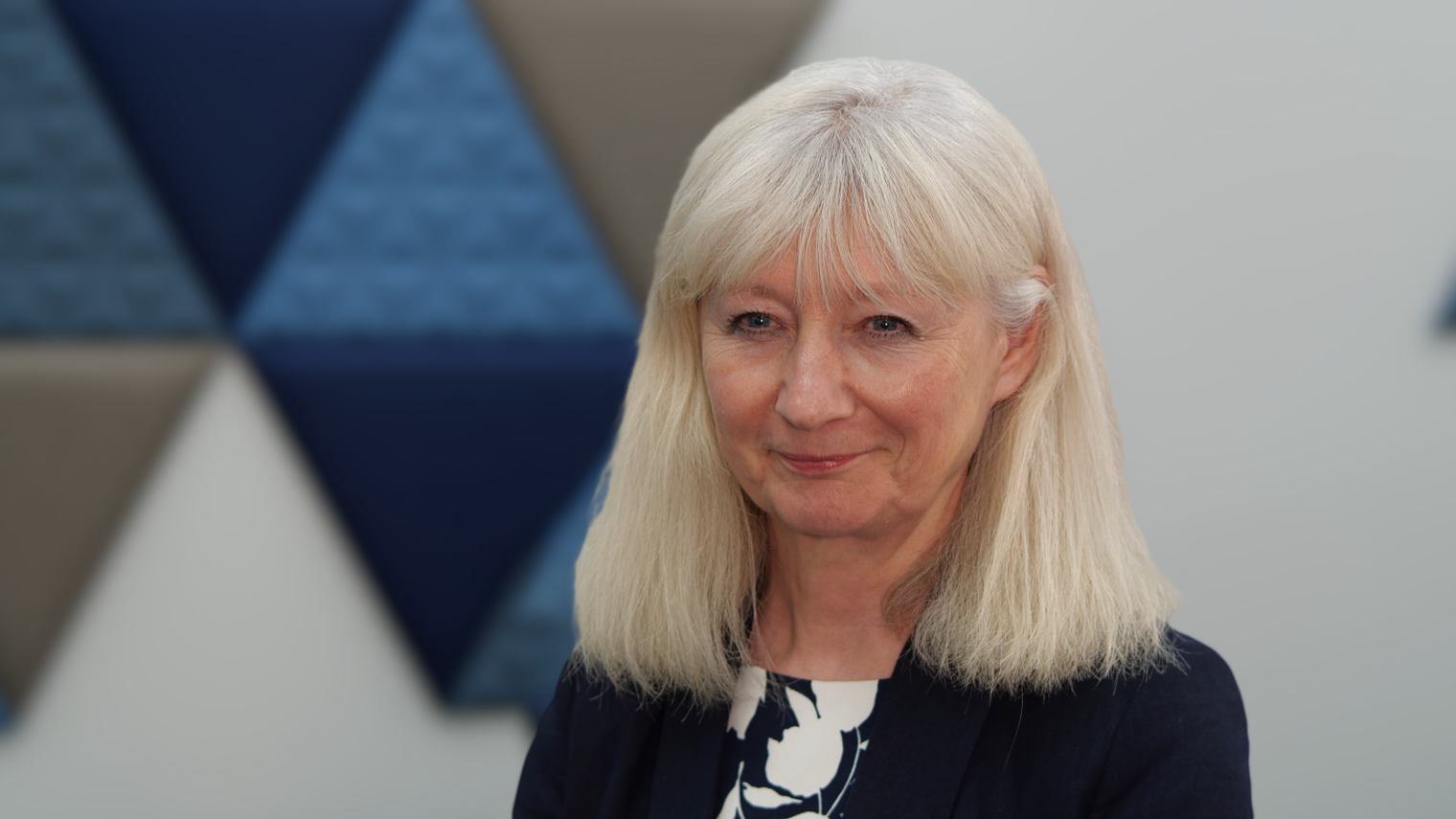 A woman with shoulder length blonde hair and wearing a black and white blouse and blazer smiles with a closed mouth