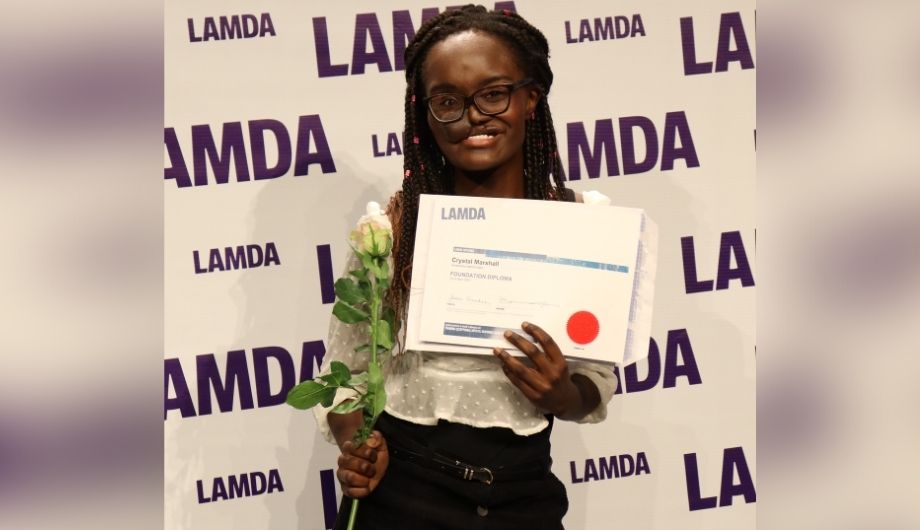 Crystal, a women in her early 20s with a skin flap. She's wearing a white blouse and black trousers and holding a rose and a graduation certificate from LAMDA. Behind her is a wall covered all over with the word 'LAMDA'