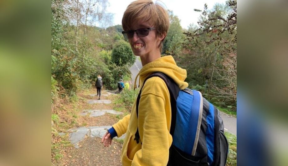 Dylan, a teenager, wearing a yellow hoodie and a blue backpack. He's outside walking down a forest path and has turned his head to smile at the camera.