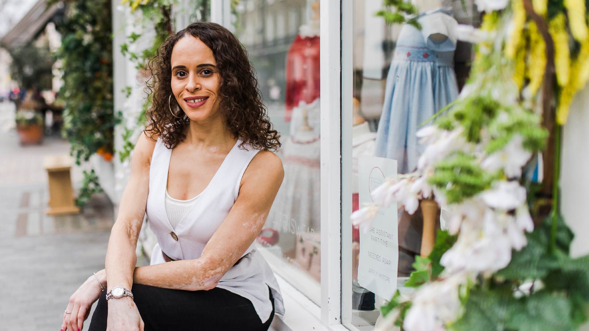 Natalie, who has vitiligo, is smiling towards the camera and wears a white vest-top and black trousers. She sits on a bench with shop windows in the background