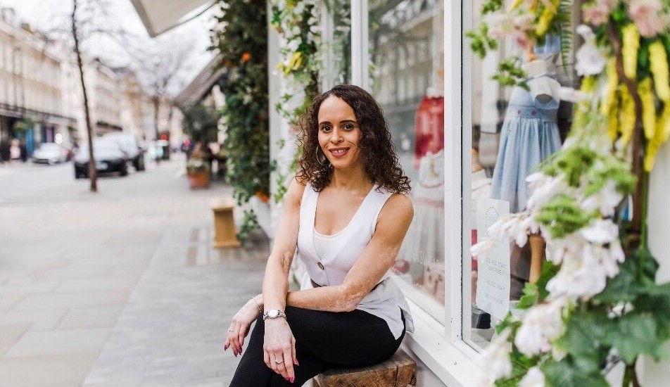A mixed race woman wearing a white vest-top and black trousers, sitting on a bench outside a shop. Patches of vitiligo are visible on her arms and hands.