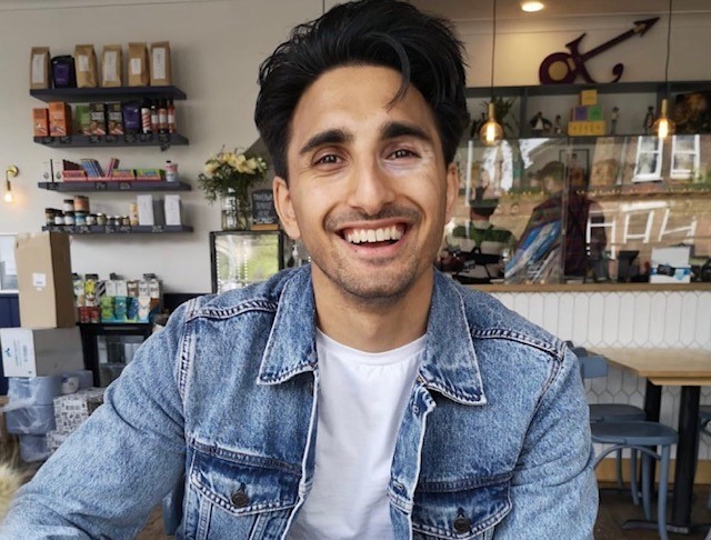 Shankar, who has vitiligo, wearing a white top and denim jacket. He is looking at the camera with a big smile. In the background are shelves with produce and a till.