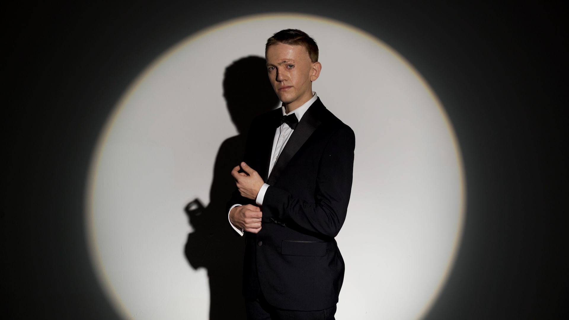 A man with a facial visible difference dressed as James Bond, in a black suit and bow tie, illuminated by a spotlight.