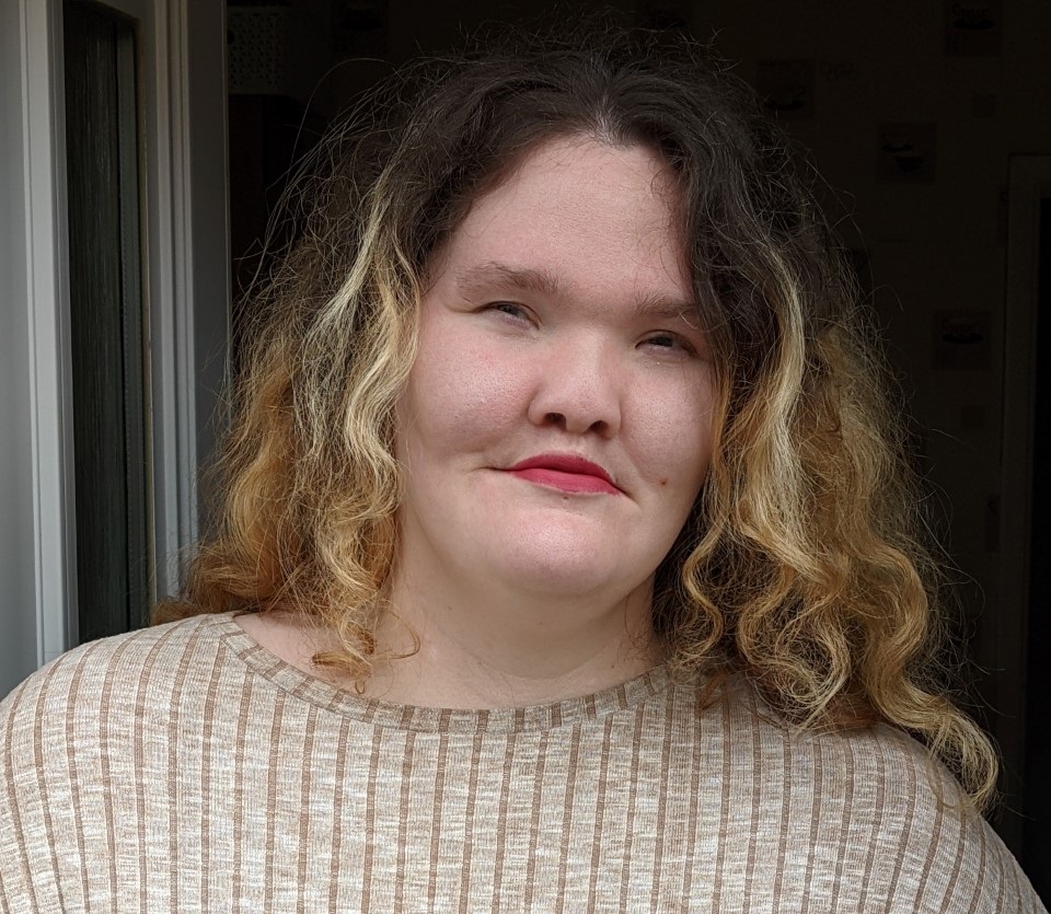 Emma, who has a facial visible difference, with wavy shoulder-length hair, wearing a beige long-sleeved top, looking at the camera