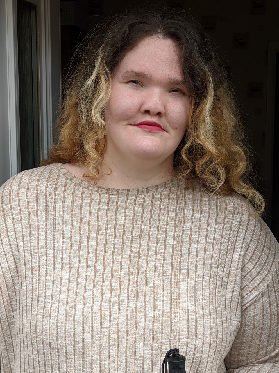 Emma, who has a facial visible difference, with wavy shoulder-length hair, wearing a beige long-sleeved top, looking at the camera