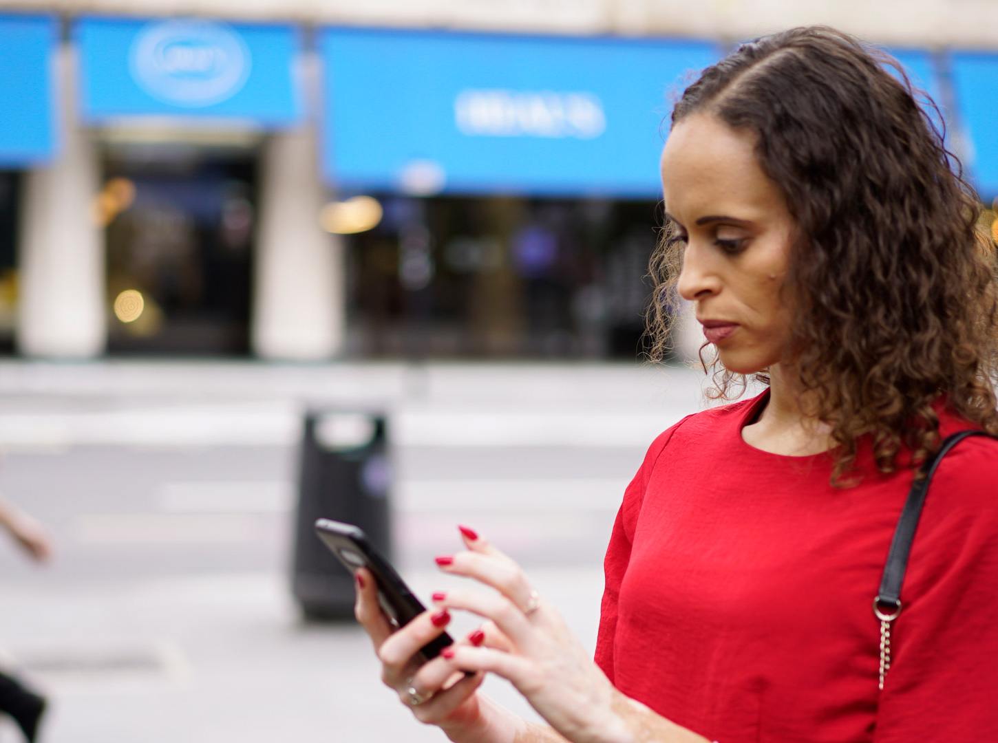 A woman with wavy, shoulder-length hair in a red top using a smartphone on the street.