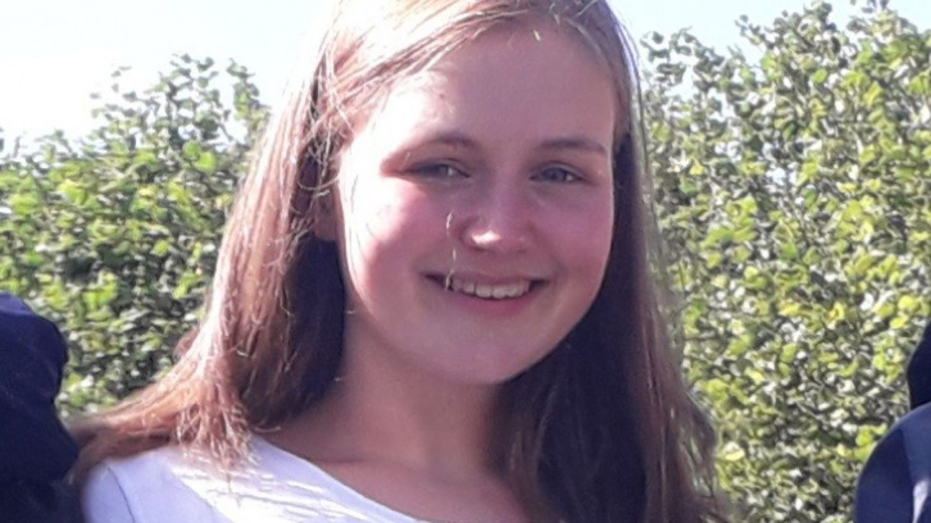 Gabby, a 17-year-old young white woman with shoulder length brown hair. She's wearing a white t-shirt, smiling, and standing in front of some green bushes.