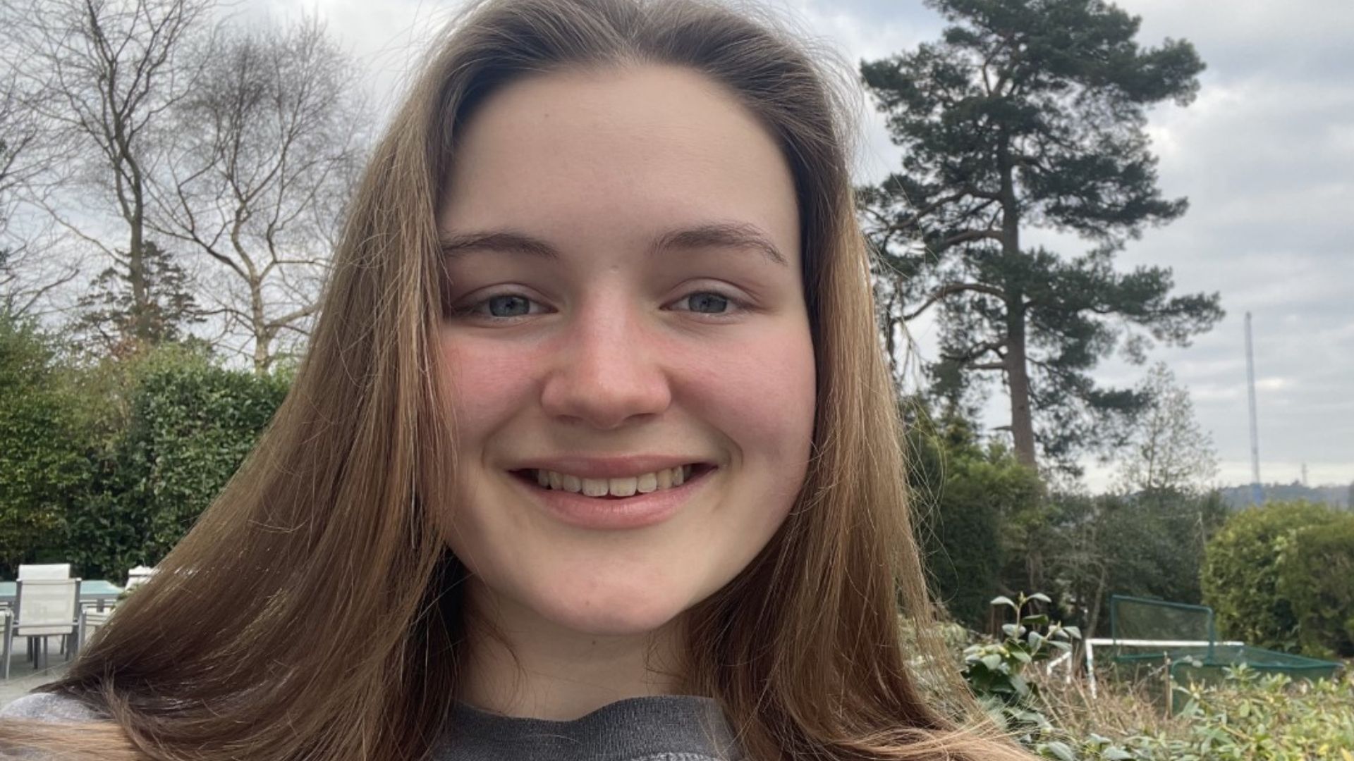 Gabby, a 17-year-old young white woman with shoulder length brown hair. She's standing outdoors in front of some trees and smiling at the camera.