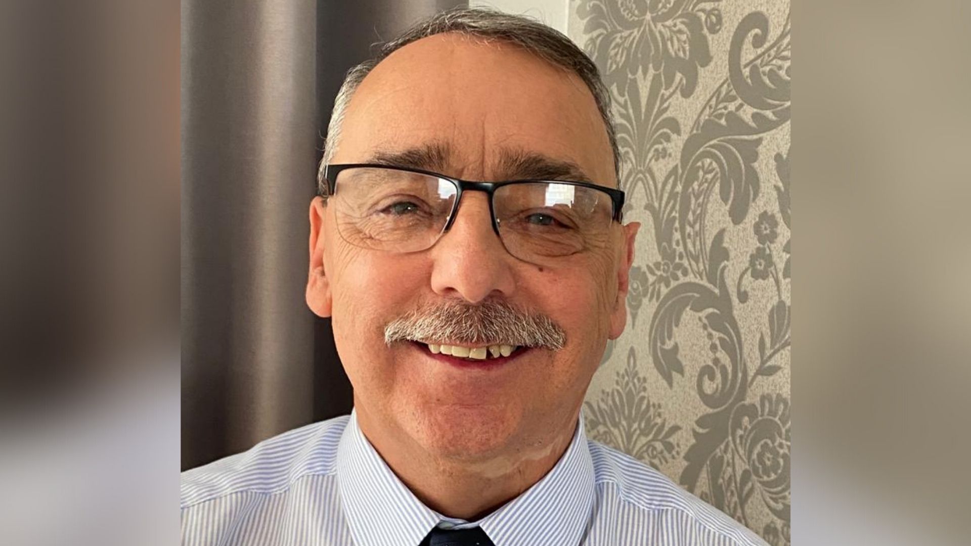 A close-up shot of Graham, a white man in his late 50s who has short grey hair and some scarring on his neck. He is wearing glasses, a blue shirt and a navy tie.