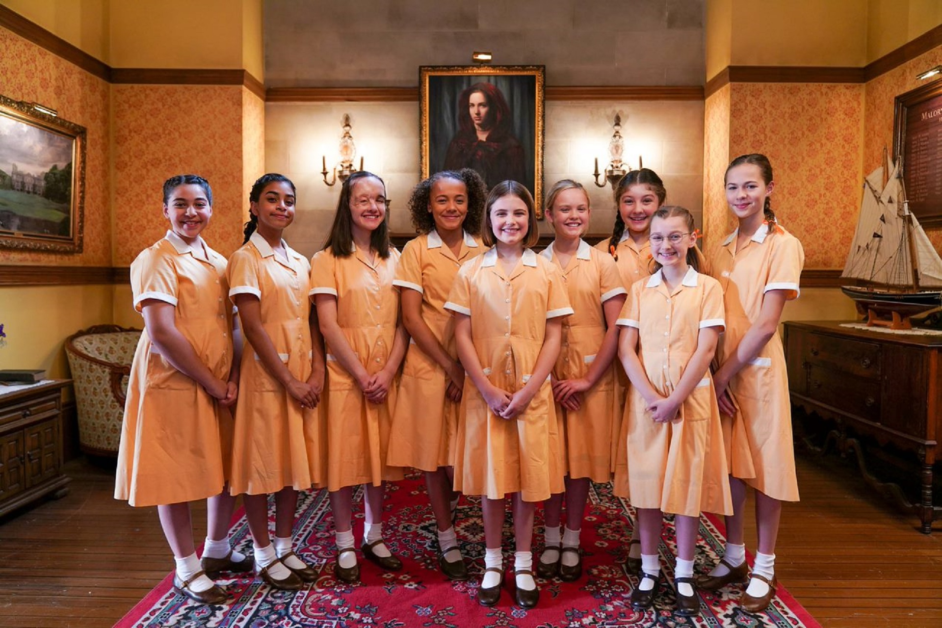 Cast of TV show Malory Towers, featuring nine senior school girls standing in their school uniform in front of a a painted portrait in a lobby.