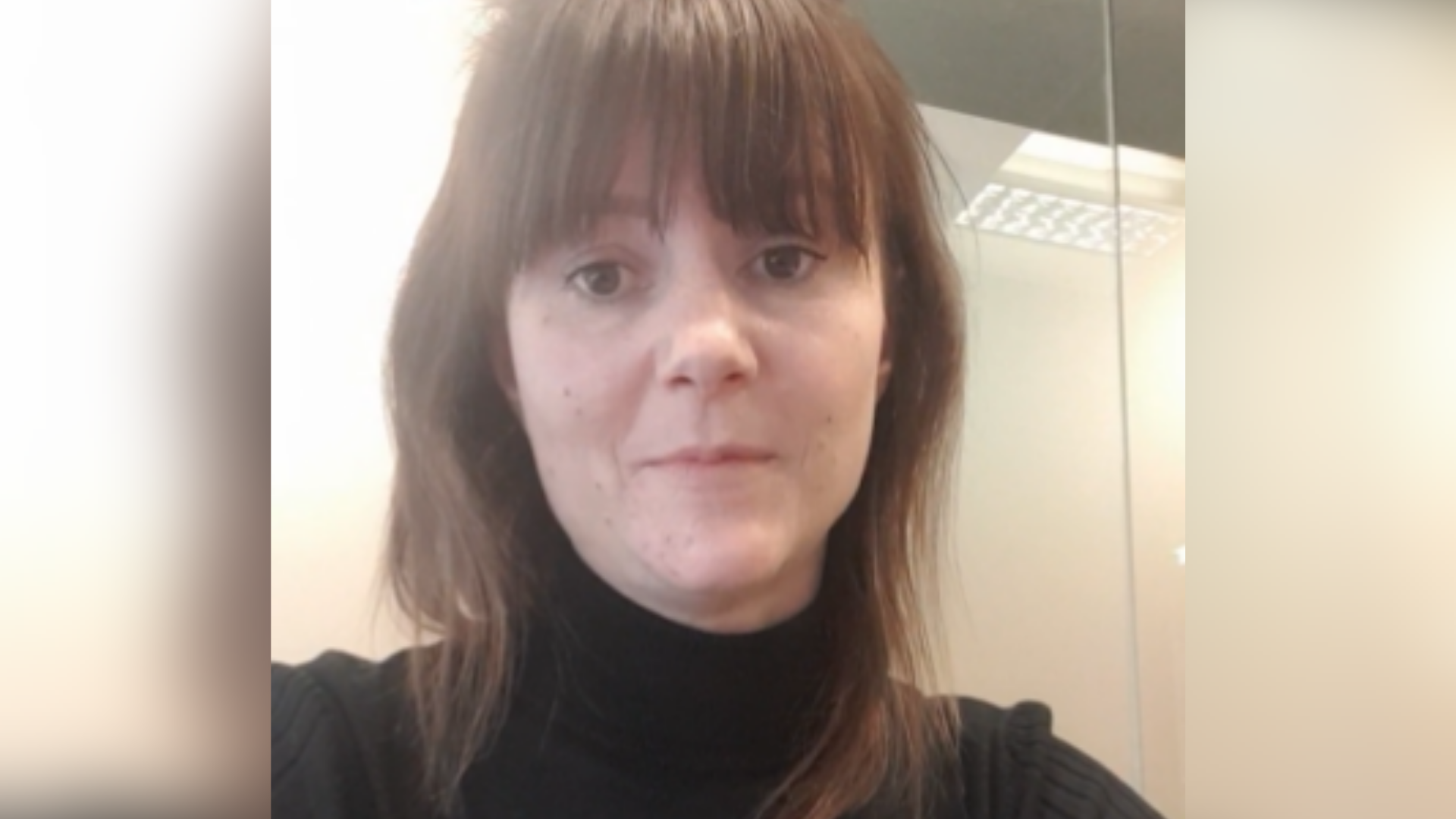 Debbie is a white woman with a long brown fringe and shoulder length brown hair. She wears a black polo neck jumper in this image.